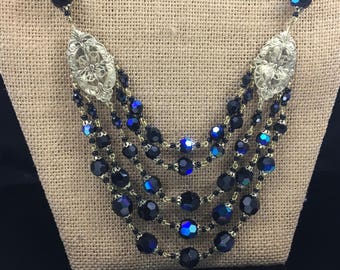 May include: A multi-strand necklace featuring black and iridescent blue beads with gold-toned metal accents. The necklace has a large, ornate pendant at each end.