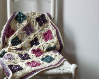 May include: A crocheted throw blanket with a white background and colorful floral designs in shades of purple, pink, teal, and green. The blanket is draped over a white wooden chair.