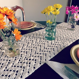 Può includere: Un runner da tavolo in crochet bianco con un motivo a chevron. Tre vasi di vetro con fiori gialli, arancioni e rosa sono disposti sul runner da tavolo. Due piatti sono sul tavolo, uno con un bordo rosso e uno con un bordo giallo.