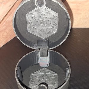 May include: A black plastic container with a hinged lid. The lid and container feature a 20-sided die design. The container is open, revealing an empty interior.
