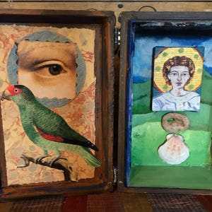 May include: A wooden box with two compartments. The left compartment features a green parrot with a red beak and a vintage image of an eye. The right compartment features a painted image of a person with a halo and a seashell.