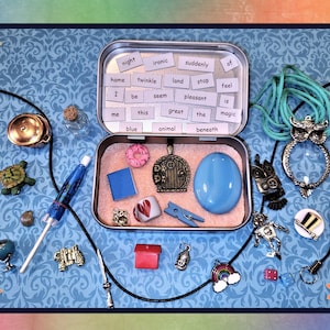 May include: A collection of small charms and trinkets, including an owl pendant, a turtle figurine, and a miniature tin with words like "magic" and "twinkle." The items are arranged on a blue patterned background with decorative flowers.