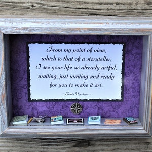 May include: A framed quote by Joni Morrison about life being a story waiting to be told. The quote is displayed on a purple background with a compass and miniature books on a shelf below.