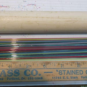 May include: A collection of colorful stained glass strips, in various colors, are displayed next to a wooden ruler and a cardboard tube. The ruler is marked with inches and the text "CLINE GLASS CO." is visible.