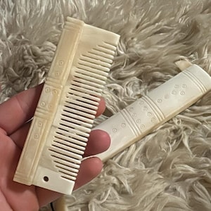 Puede incluir: Dos peines blancos hechos de hueso o marfil. El peine de la izquierda tiene un patrón decorativo y se sostiene en una mano. El peine de la derecha está sobre una superficie blanca y esponjosa.