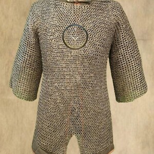 May include: A chainmail shirt with a high collar and short sleeves. The shirt is made of small, interlocking metal rings. The collar is a darker color with a leather strap and buckle. The shirt extends to mid-thigh and has a split at the bottom.