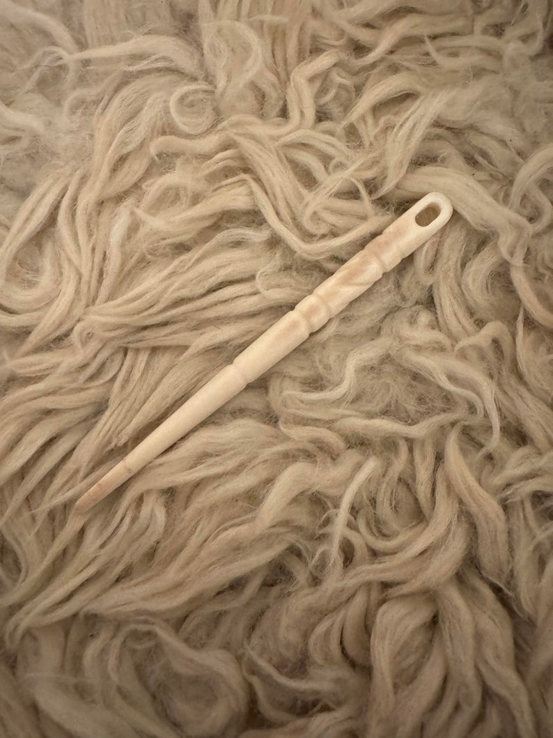 Puede incluir: Una aguja de madera blanca con un bucle en la parte superior, que se encuentra sobre un lecho de fibras de lana beige.