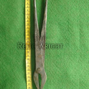 May include: A pair of antique metal tongs, approximately 32 inches long, with a curved, pointed end. The tongs are a dark, aged metal, and rest on a green surface. A yellow measuring tape is placed next to the tongs.