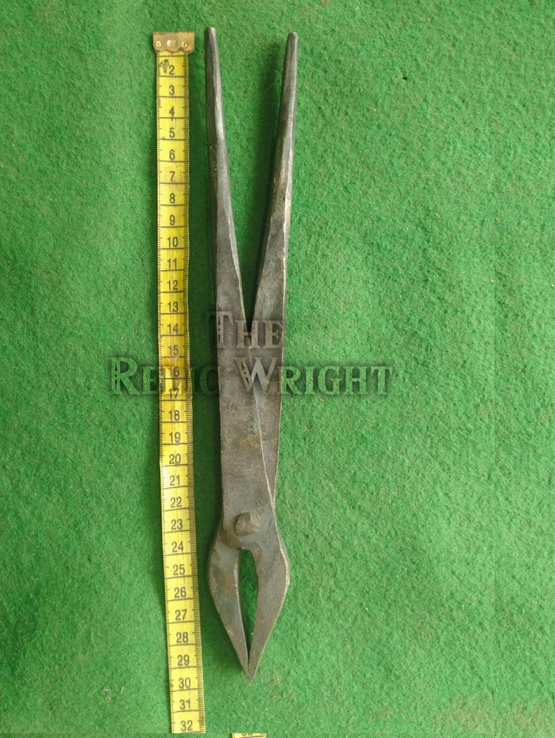 May include: A pair of antique metal tongs, approximately 32 inches long, with a curved, pointed end. The tongs are a dark, aged metal, and rest on a green surface. A yellow measuring tape is placed next to the tongs.