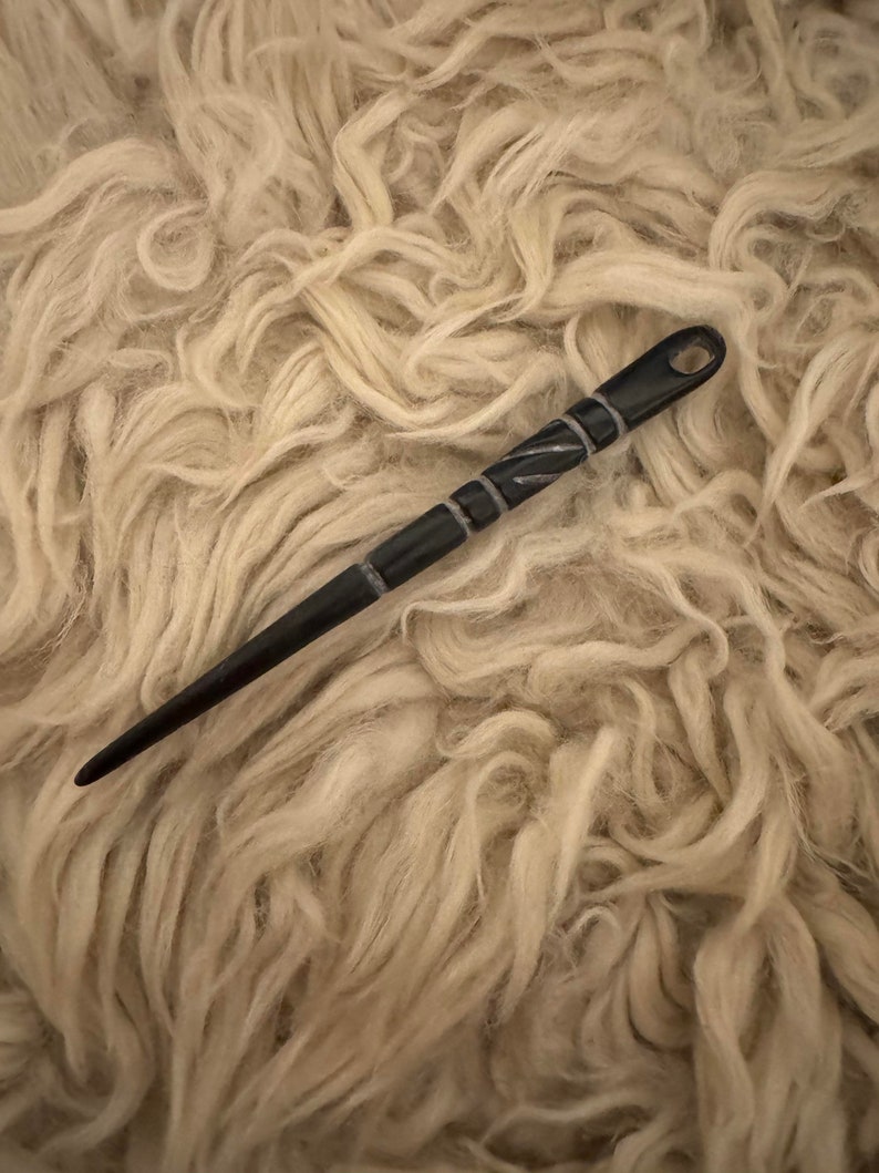 Puede incluir: Una aguja de madera negra con un dise&ntilde;o tallado, que se encuentra sobre una superficie blanca y esponjosa.
