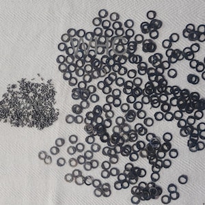 Op de afbeelding: Een grote stapel zwarte metalen ringen op een wit stoffen oppervlak. Links van de ringen ligt een kleinere stapel zwarte metalen onderdelen.