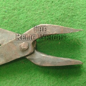 May include: Close-up of antique metal tool with the text "THE RELIC WRIGHT" stamped on the hinge. The tool has a rusty, aged appearance, with pointed ends and a central pivot. The background is a textured green surface.