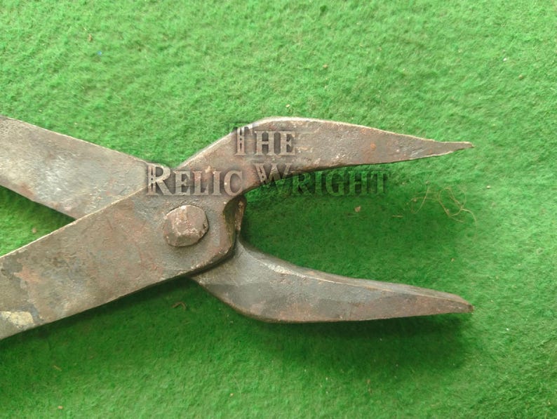 May include: Close-up of antique metal tool with the text "THE RELIC WRIGHT" stamped on the hinge. The tool has a rusty, aged appearance, with pointed ends and a central pivot. The background is a textured green surface.