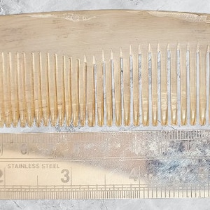Puede incluir: Un peine de madera con mango redondeado y dientes finos. El peine mide aproximadamente 18 cm de largo y se muestra junto a una regla para la escala.