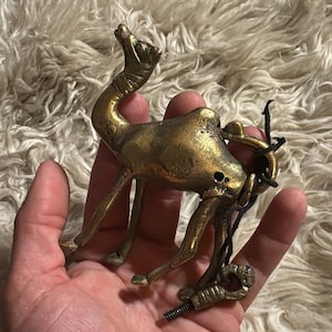 May include: A gold-colored metal camel figurine with a black cord attached to the back. The camel is standing on a white fluffy surface.