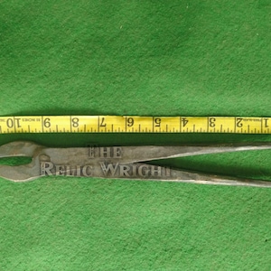 May include: A pair of metal tongs with the text "THE RELIC WRIGHT" etched on the handle. A yellow measuring tape shows the tongs are approximately 11 inches long. The tongs are on a green surface.