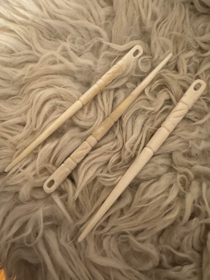 Puede incluir: Tres agujas de tejer de hueso con tallas intrincadas. Las agujas est&aacute;n sobre una superficie esponjosa de color crema.