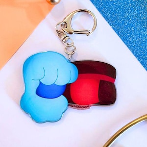 May include: A keychain featuring a cartoon character design. The keychain has a blue cloud-like shape and a red and brown shape, connected by a silver metal ring and clasp. The background is a mix of white, blue, and peach.