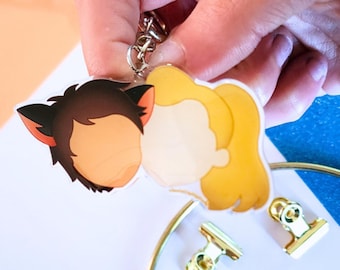 May include: A clear acrylic keychain featuring two cartoon characters, one with brown hair and cat ears, the other with blonde hair. The keychain is attached to a silver metal keyring. Gold-colored binder clips and a gold-colored ring are in the background.