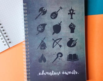 May include: Spiral-bound notebook with a dark blue and gray cover featuring various black icons, including a shield, bow and arrow, and a book. The words ".adventure awaits." are printed at the bottom.