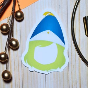 Puede incluir: Una pegatina troquelada con un diseño de gnomo caprichoso. El gnomo tiene un sombrero azul con un acento amarillo, una cara blanca y una barba verde claro. La pegatina está sobre una superficie de madera con elementos decorativos dorados.