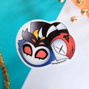 May include: A die-cut sticker featuring two cartoon characters. One character is blue with red eyes, and the other is red and white with an X for an eye. The sticker has the text "@wingingitstudio".