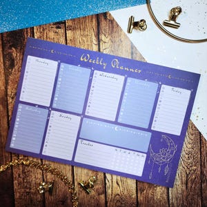 May include: A purple weekly planner with a gold "Weekly Planner" title. The planner has sections for each day of the week, a tracker, and a moon and dreamcatcher design. The planner is on a wooden surface.
