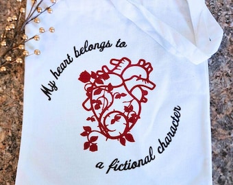 May include: White tote bag with the text "My heart belongs to a fictional character" in black script. A red anatomical heart design with floral accents is centered on the bag. The bag has white handles.