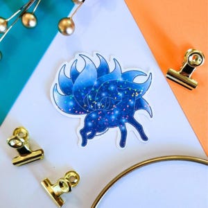 May include: A sticker featuring a blue and white nine-tailed fox design with a starry night sky pattern. The sticker is on a white surface with gold binder clips and a gold ring. The background is teal and orange.