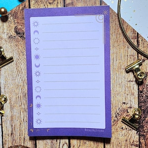 May include: A purple notepad with a celestial design featuring suns, moons, and stars. The notepad has lined pages and a gold border. The text "@WINGINGITSTUDIO" is printed on the bottom right corner.