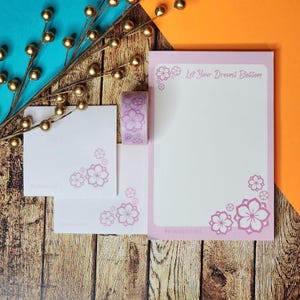 Puede incluir: Un juego de papelería con un bloc de notas, notas adhesivas y cinta washi. Los artículos son blancos con detalles florales rosas y el texto "Let Your Dreams Blossom". El conjunto está dispuesto sobre una superficie de madera.
