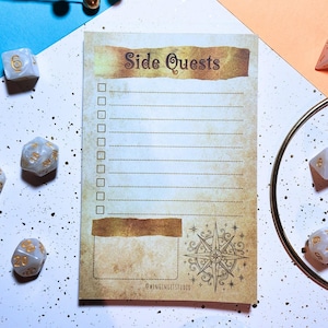 May include: A notepad with the words "Side Quests" in a vintage font. The notepad has a checklist design with a compass rose graphic and the text "@wingingitstudio". Several dice are scattered around the notepad.