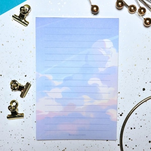 May include: A lined notepad with a sky-themed design featuring clouds in shades of blue, pink, and white. The notepad has a light blue background and is surrounded by gold binder clips and decorative pins. The text "@walkinglittlestudio" is at the bottom.