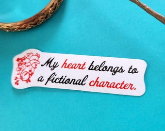 May include: White sticker with the text "My heart belongs to a fictional character" in black and red. The sticker has a red heart and floral design on the left side. A gold bracelet is in the upper left corner.