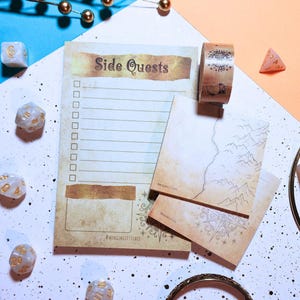 May include: A notepad with the words "Side Quests" and a checklist, accompanied by a roll of decorative tape and small map cards. The items are arranged on a white and blue surface, with dice scattered around.