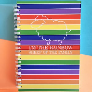 以下が含まれることがあります： 虹色のストライプの表紙と白い羊の輪郭が特徴の、スパイラル綴じノート。表紙には「I'M THE RAINBOW SHEEP OF THE FAMILY」の文字が印刷されています。カラフルな背景にノートが置かれています。