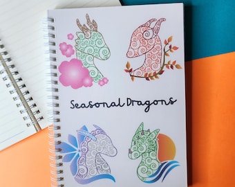 Może przedstawiać: Spiralny notatnik z napisem "Seasonal Dragons" na okładce. Okładka zawiera cztery ilustracje smoków w kolorach zielonym, czerwonym, białym i niebieskim, z unikalnymi elementami sezonowymi.