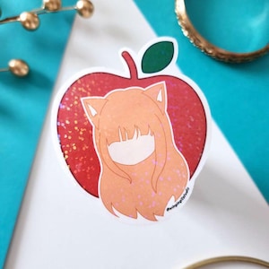 以下が含まれることがあります： 赤いリンゴのステッカー。キラキラした質感で、猫耳とオレンジ色の髪の毛を持つ漫画のキャラクターが描かれています。キャラクターの顔は白く、ステッカーには白い縁があります。ステッカーは白と青の表面にあります。