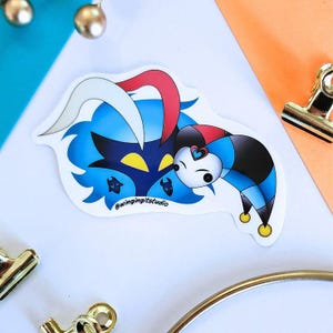 May include: A die-cut sticker featuring a cartoon illustration of a blue demon-like character with yellow eyes and a jester character. The jester has a white face, a heart, and a black, red, and blue outfit. The sticker has the text "@wingingitstudio".