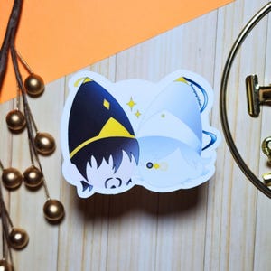 Puede incluir: Una pegatina con dos personajes de dibujos animados con pelo blanco y negro, que llevan sombreros con detalles amarillos. Un personaje tiene un sombrero negro, el otro un sombrero blanco. La pegatina tiene un borde blanco y está sobre una superficie de madera.