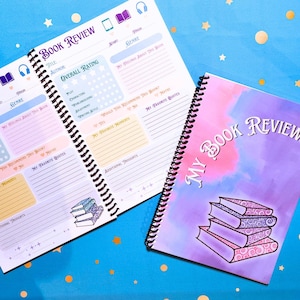 May include: Two spiral-bound book review journals. One is open, displaying lined pages with prompts for title, author, and overall rating. The other journal has a purple cover with the text "My Book Review" and a stack of books.