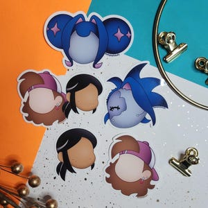 May include: A collection of colorful stickers featuring cartoon characters with various hairstyles and accessories. The stickers include characters with blue hair, pink hats, and black hair. The stickers are on a white background with gold accents.
