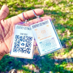 May include: Two clear acrylic keychains with silver rings are held up against a blurred green background. Each keychain has a QR code and the text "JOURNEY BODYWORKS". One keychain says "LINKTREE" and the other has social media icons.