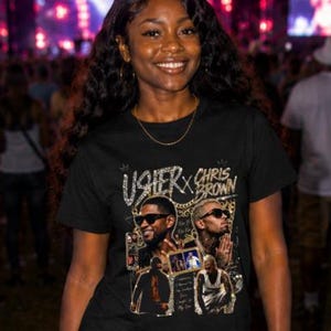 May include: A black short-sleeve t-shirt featuring a graphic print of Usher and Chris Brown with the text Usher x Chris Brown. The shirt has a crew neckline and is paired with black leather-like pants, creating a dark, coordinated outfit.