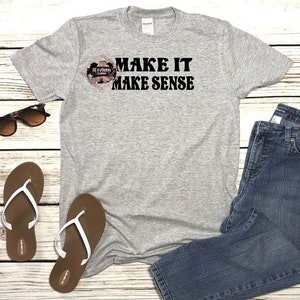May include: A gray t-shirt with the text "MAKE IT MAKE SENSE" printed in black. The shirt is laid flat on a wooden surface with a pair of sunglasses, a pair of brown sandals, and a pair of blue jeans.