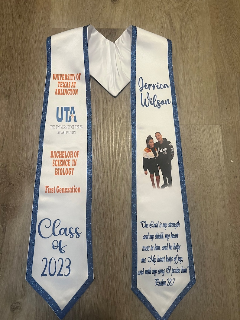 May include: White graduation stole with blue trim and glitter. The stole features the University of Texas at Arlington logo, the text "Bachelor of Science in Biology", "First Generation", "Class of 2023", and a photo of a person with the name "Jerrica Wilson". The stole also includes the text "The Lord is my strength and my shield, my heart trusts in him, and he helps me. My heart leaps of joy, and with my song I praise him." Psalm 28:7