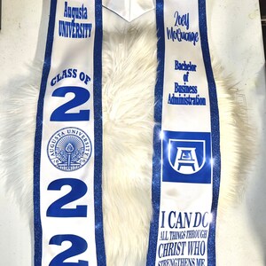 May include: White graduation stole with blue trim and glitter. The stole features the Augusta University logo, the text "Class of 2022", and the graduate's name "Zoey Moluaige". The stole also includes the text "Bachelor of Business Administration" and the quote "I can do all things through Christ who strengthens me - Philippians 4:13".