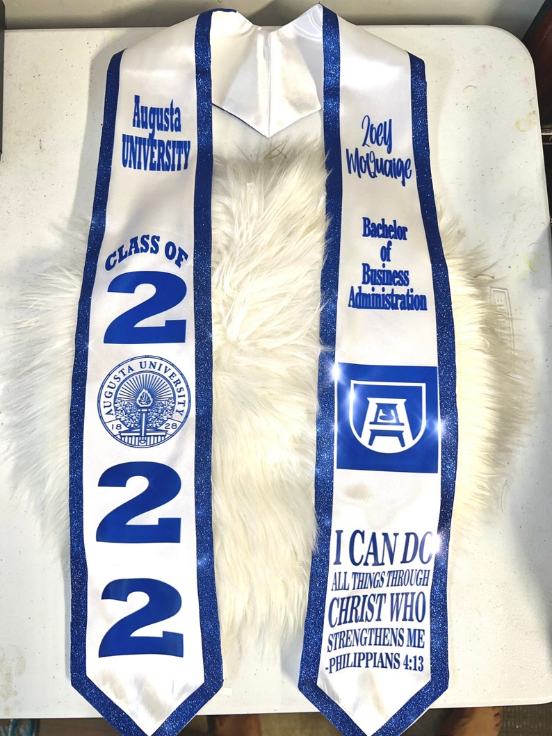 May include: White graduation stole with blue trim and glitter. The stole features the Augusta University logo, the text "Class of 2022", and the graduate's name "Zoey Moluaige". The stole also includes the text "Bachelor of Business Administration" and the quote "I can do all things through Christ who strengthens me - Philippians 4:13".