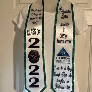 May include: White graduation sash with green trim and glitter. The sash says "Florida State College at Jacksonville "FSCJ" Class of 2022" on one side and "Cassandra Lewis Associate in Science Funeral Service" on the other side. The sash also has a small logo of the college.
