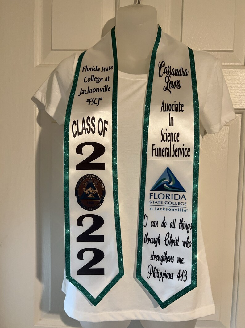 May include: White graduation sash with green trim and glitter. The sash says "Florida State College at Jacksonville "FSCJ" Class of 2022" on one side and "Cassandra Lewis Associate in Science Funeral Service" on the other side. The sash also has a small logo of the college.